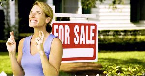 Establishing asking price. Woman with fingers crossed standing in from of home for sale sign.